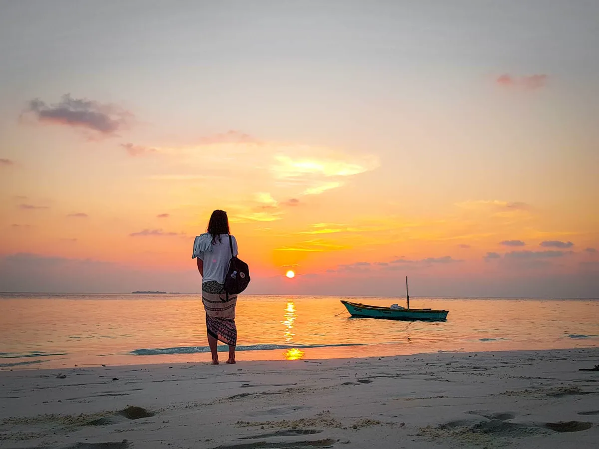 Woman standing at the water's edge watching a golden sunset on a Maldivian beach, with a traditional dhoni visible offshore
